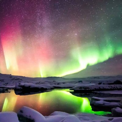 Colorful Northern Lights Dancing Over a Lake With Snow Covered Rocks in Iceland