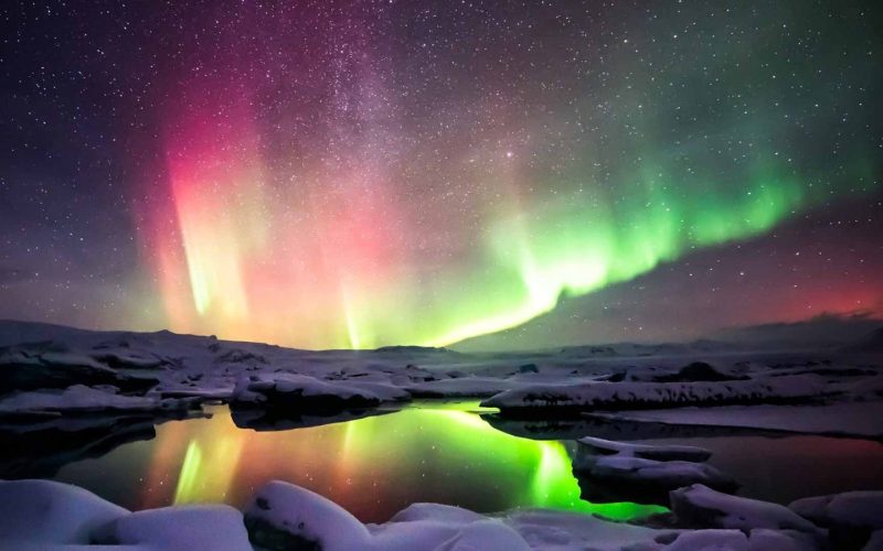 Colorful Northern Lights Dancing Over a Lake With Snow Covered Rocks in Iceland