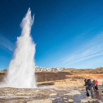 People Standing By Erupting Strokkur In The Great Geysir Hot Springs Area On The Golden Circle In Iceland