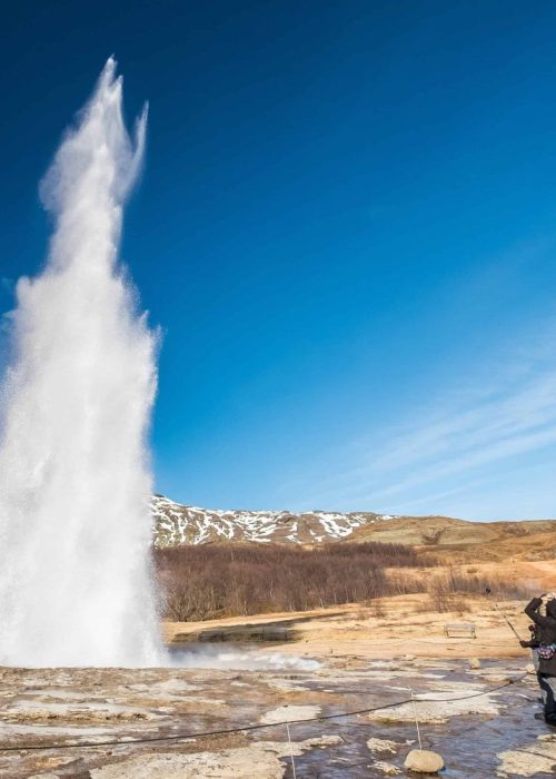 People Standing By Erupting Strokkur In The Great Geysir Hot Springs Area On The Golden Circle In Iceland