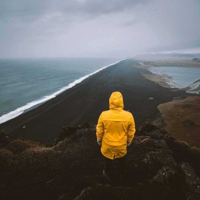 Man Wearing a Yellow Raincoat Standing on the Edge of a Cliff over a Black Sand Beach in South Iceland