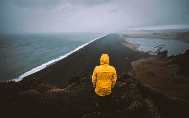 Man Wearing a Yellow Raincoat Standing on the Edge of a Cliff over a Black Sand Beach in South Iceland